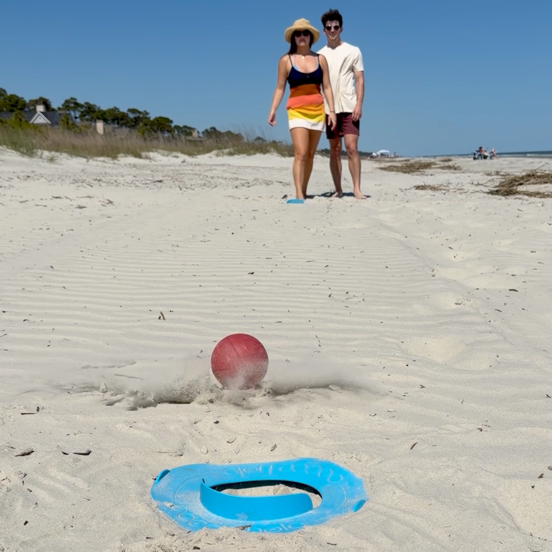 Two people on a sandy beach playing the best new beach game of 2026 - bocce hole.  the bocce ball has been tossed on soft sand and is rolling towards the scoring ring