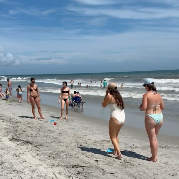 People on a beach with a clear sky playing  the best beach game of 2026 - bocce hole beach game.  four girls are playing, and one rolled a ball carefully hard sand