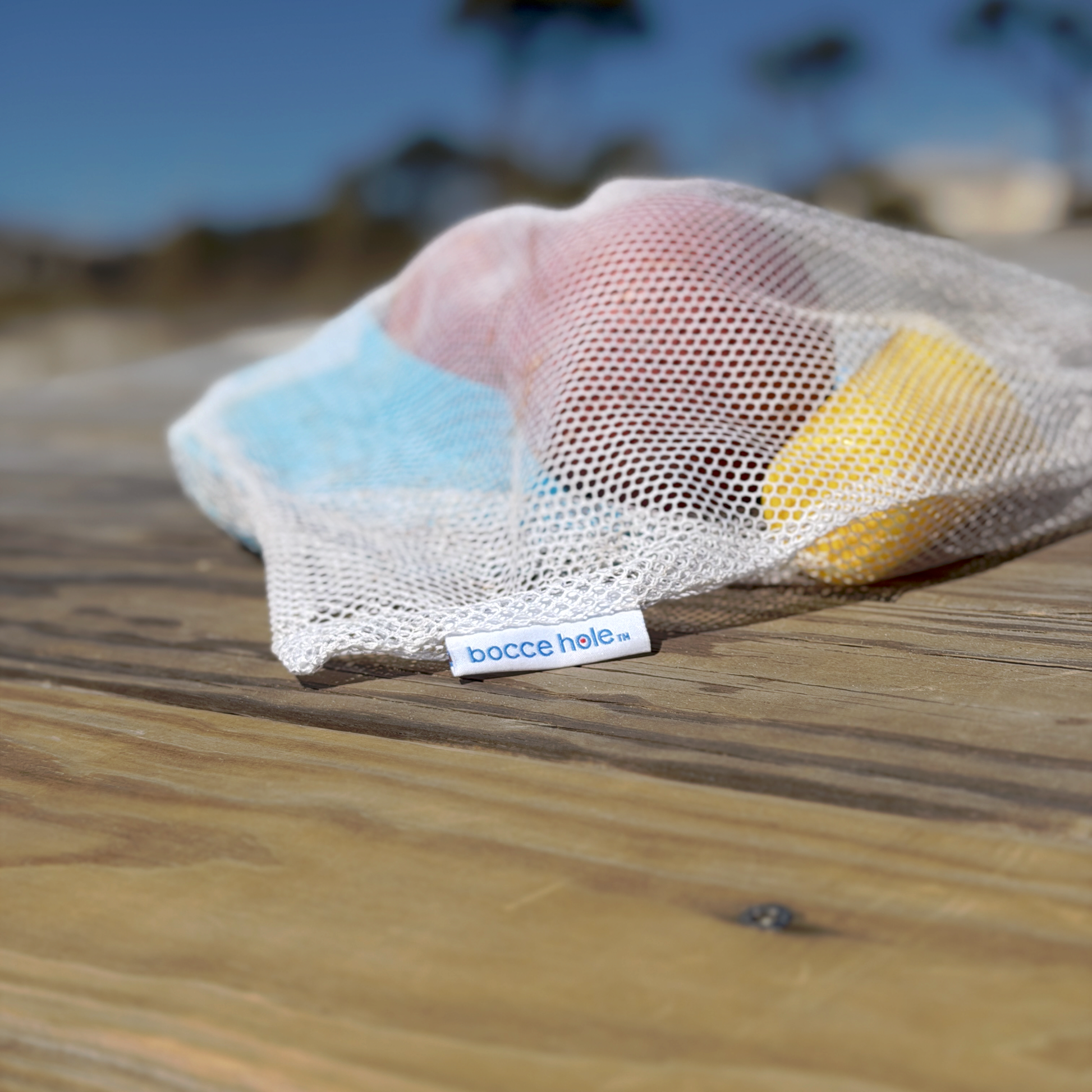 Bocce hole set with two balls, two sand scoops, and two rings in the branded mesh bag.  The bag is laying on a wooden beachside picnic table, ready for another round of play.   