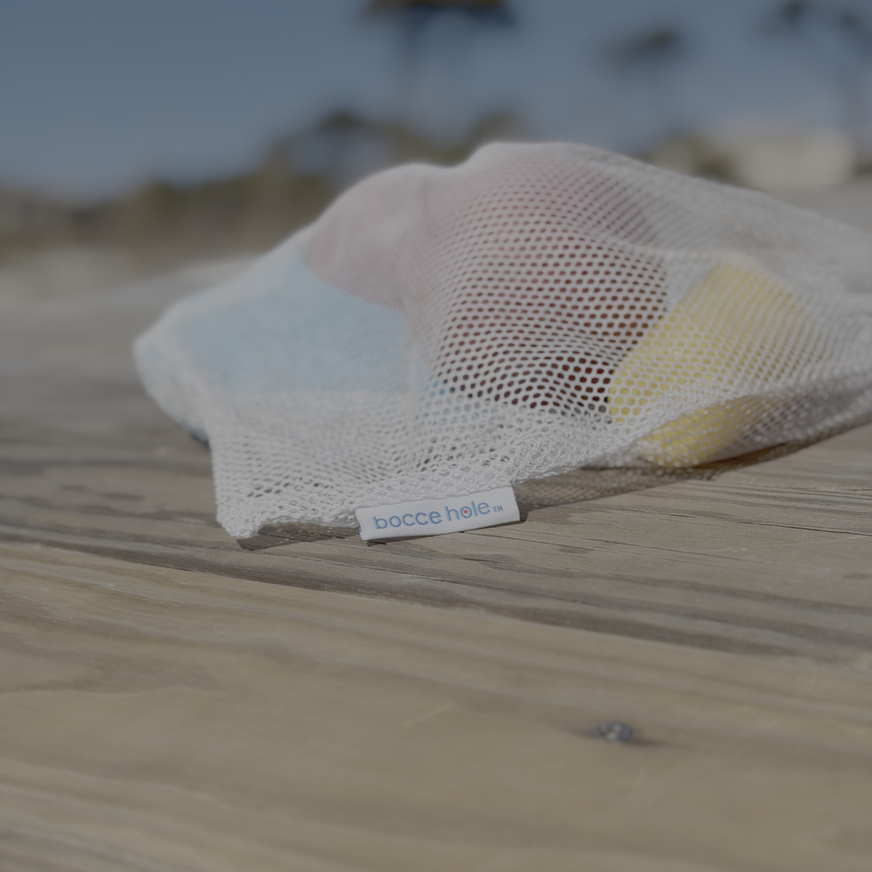 Bocce hole set with two balls, two sand scoops, and two rings in the branded mesh bag.  The bag is laying on a wooden beachside picnic table, ready for another round of play.   